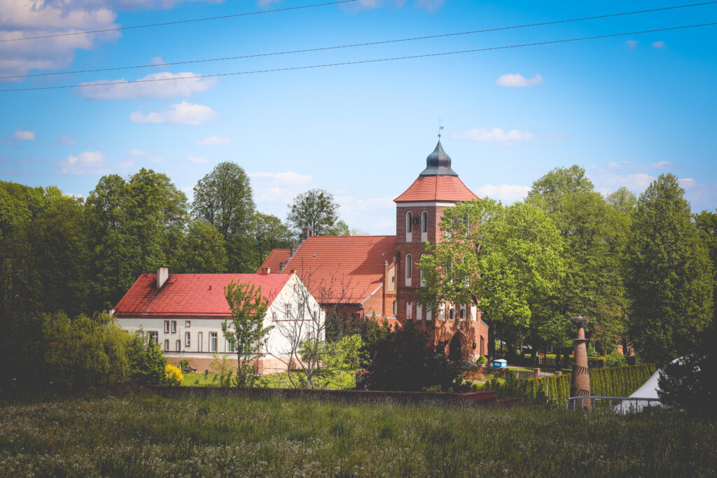 Widok na zabytkowy budynek z czerwoną dachówką otoczony zielenią.