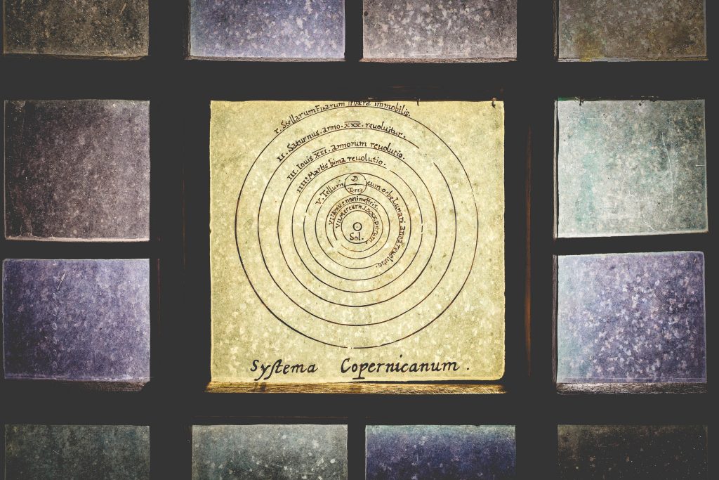 Ilustracja przedstawia diagram układu słonecznego według teorii Mikołaja Kopernika, z oznaczeniem Słońca w centrum i okręgami symbolizującymi orbity planet. Na żółtej kartce znajduje się napis "Systema Copernicum". Tło stanowią kolorowe, stłuczone szyby.