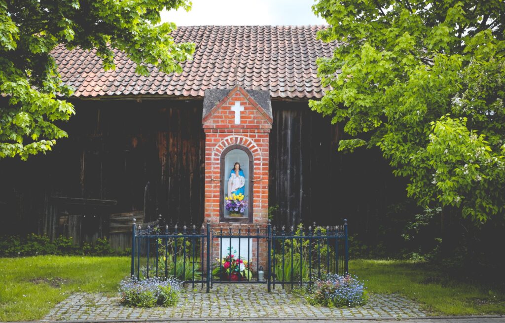 Kapliczka z figurą Maryi, otoczona zielenią.