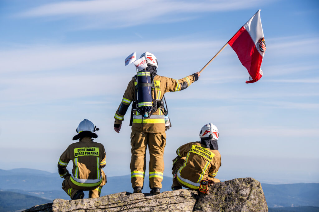 Strażacy stoją na szczycie góry, jeden trzyma flagę.