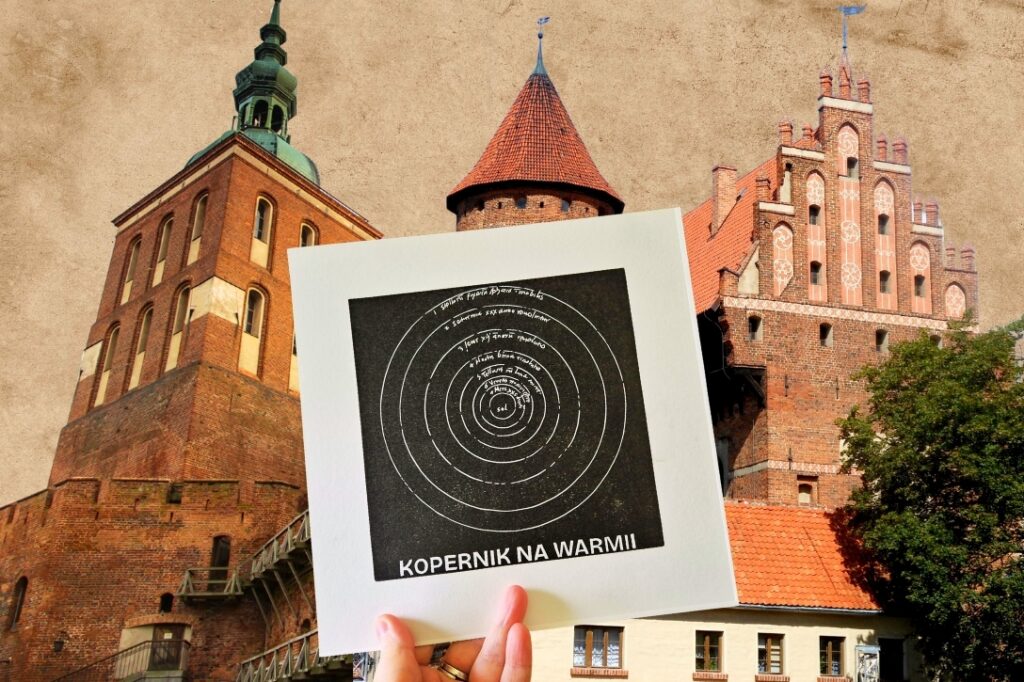 Zbliżenie na grafikę z napisem 'Kopernik na Warmii' przed zabytkowymi budynkami.
