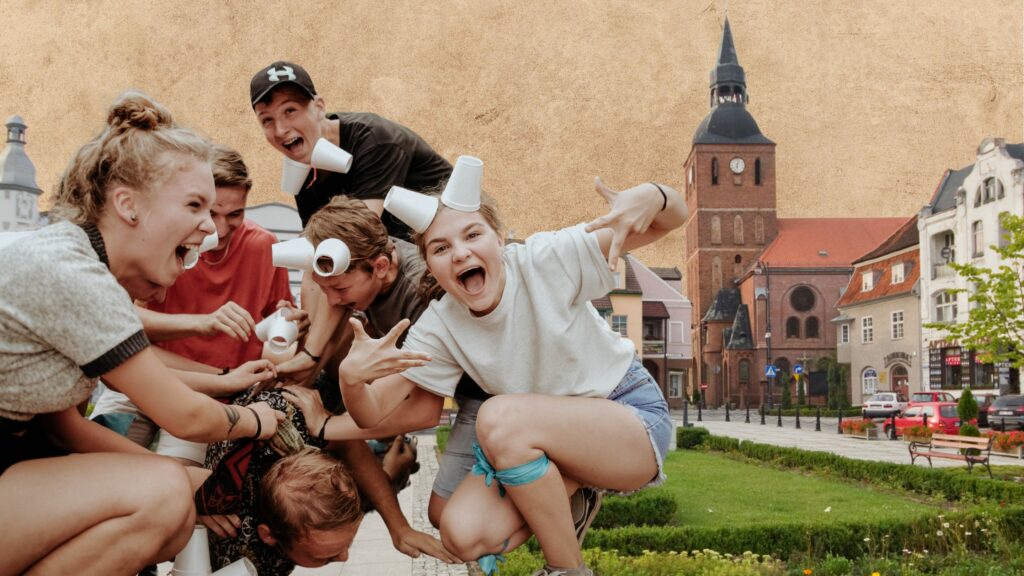 Grupa młodzieży bawi się na ulicy, niektórzy mają kubki na głowach.