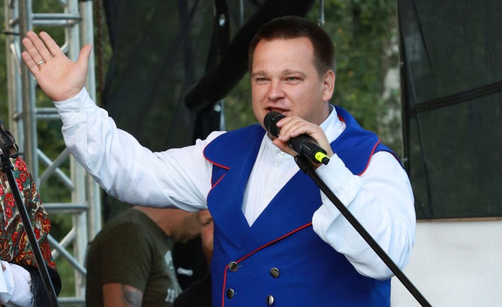 Mężczyzna w niebieskiej kamizelce śpiewa do mikrofonu na scenie.