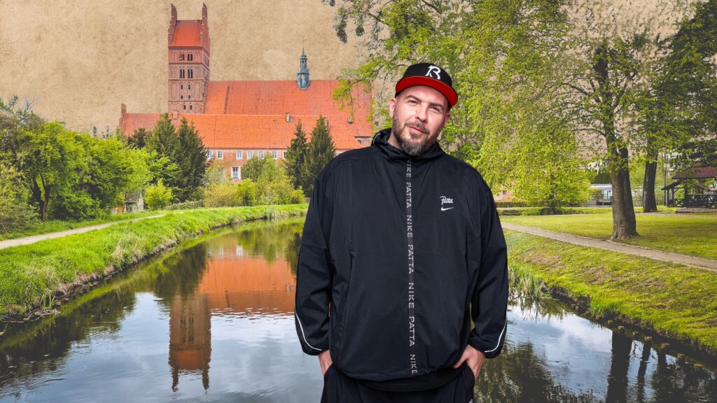 Mężczyzna w czarnej kurtce na tle rzeki i budynków.