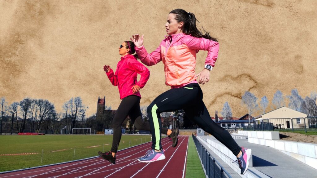 Dwie biegaczki w kolorowych kurtkach na torze lekkoatletycznym.