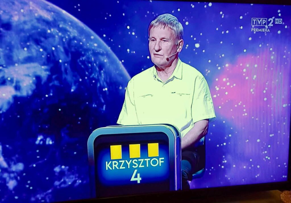 Mężczyzna siedzący na scenie telewizyjnej z kosmicznym tłem.