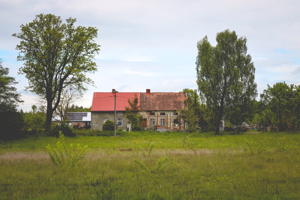 Stary dom z czerwoną dachówką w otoczeniu drzew i trawy.