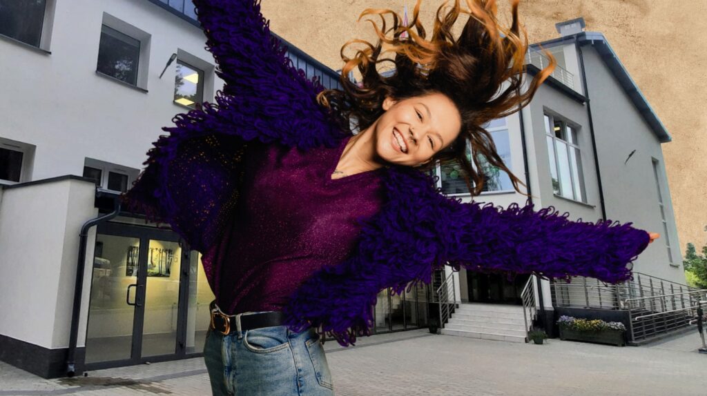 Uśmiechnięta dziewczyna w kolorowym swetrze skacze na tle budynku.