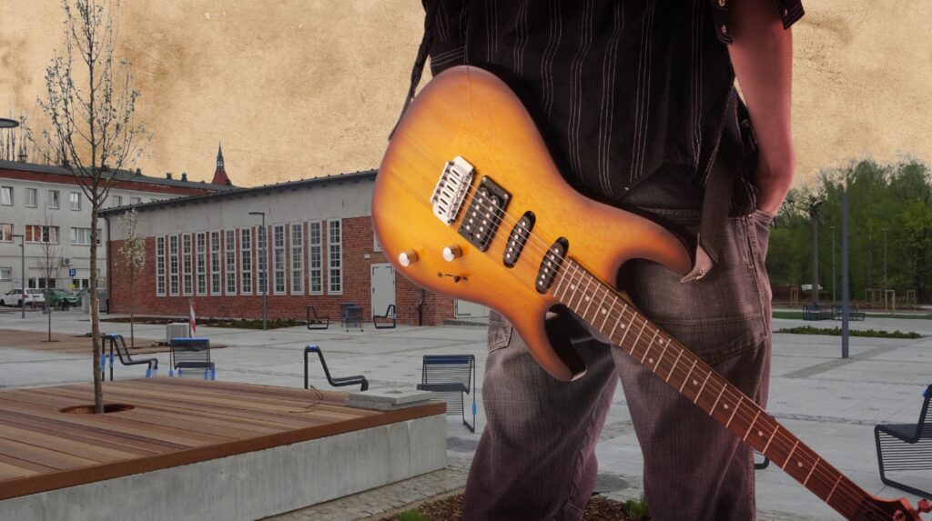 Osoba trzymająca gitarę elektryczną na tle budynku.