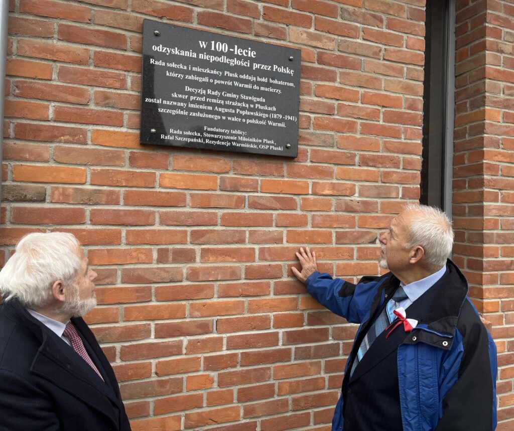 Dwóch mężczyzn ogląda tablicę pamiątkową na ceglanej ścianie.