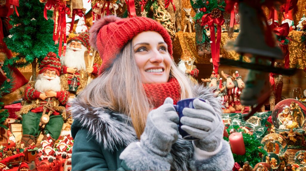 Kobieta w czerwonym czapce trzyma filiżankę z gorącym napojem przed ozdobami świątecznymi.