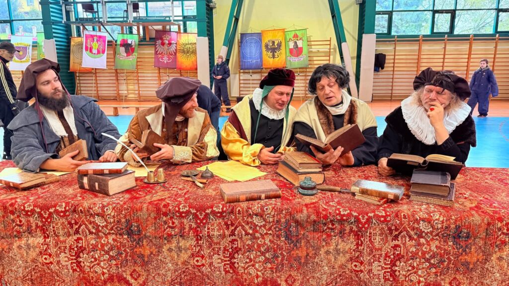 Grupa mężczyzn w historycznych strojach przegląda książki przy stole.