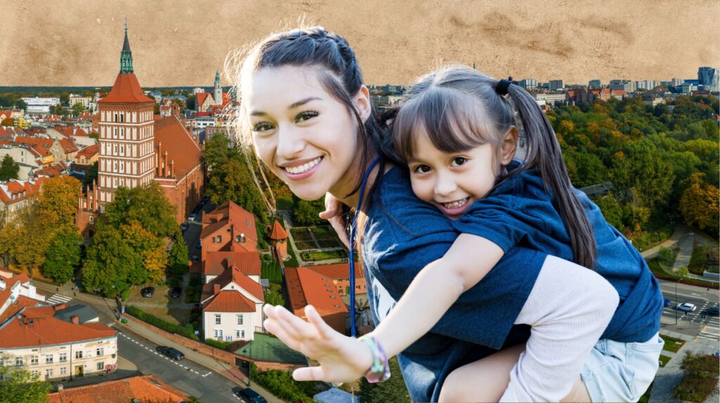 Młoda kobieta niesie na plecach dziewczynkę z uśmiechem na tle miasta.