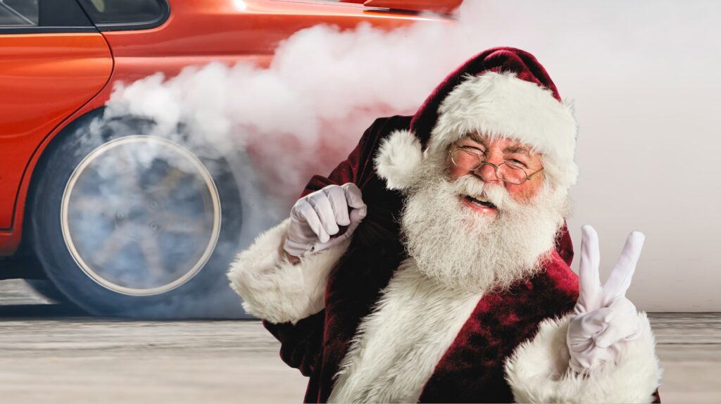 Święty Mikołaj robi znak pokoju obok palącego się samochodu.