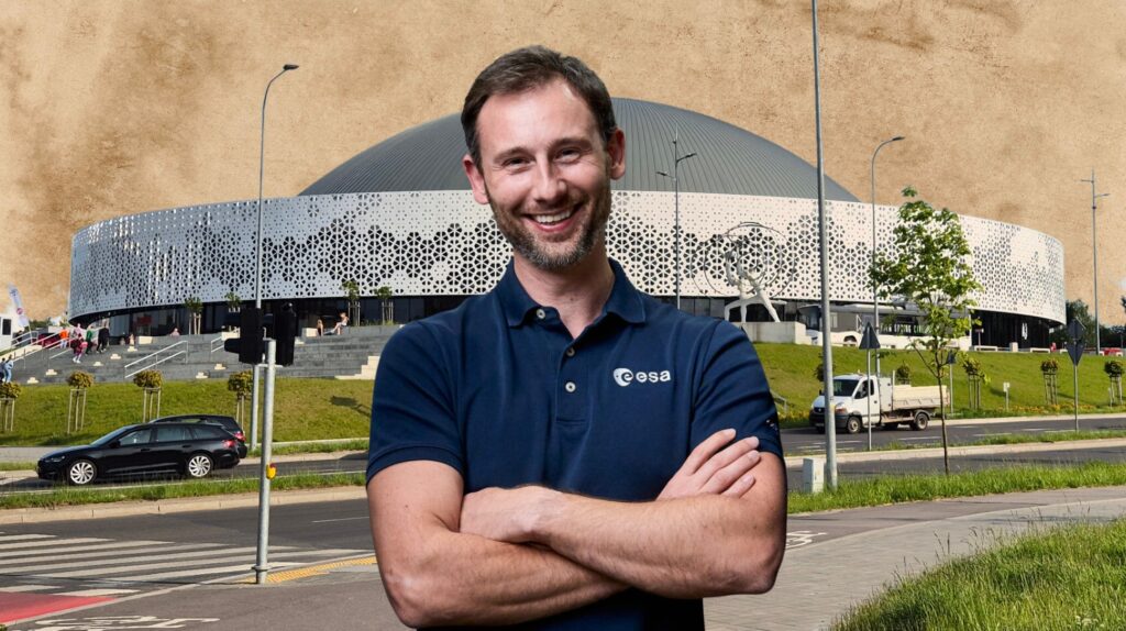 Mężczyzna w koszulce ESA z uśmiechem przed nowoczesnym budynkiem.