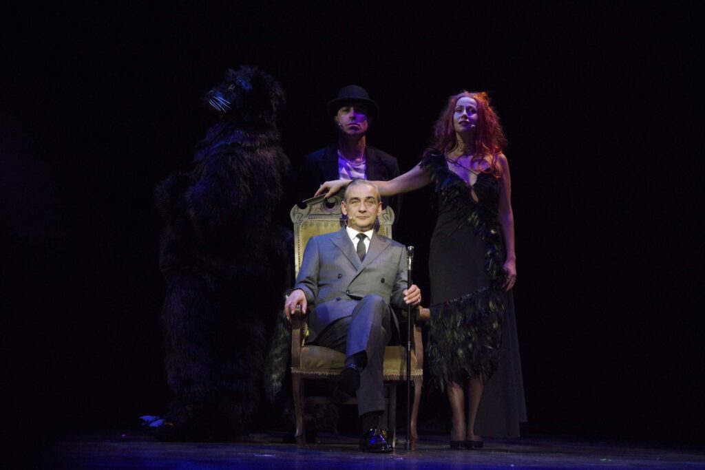 Muzyk w eleganckim garniturze na scenie z dwiema postaciami.