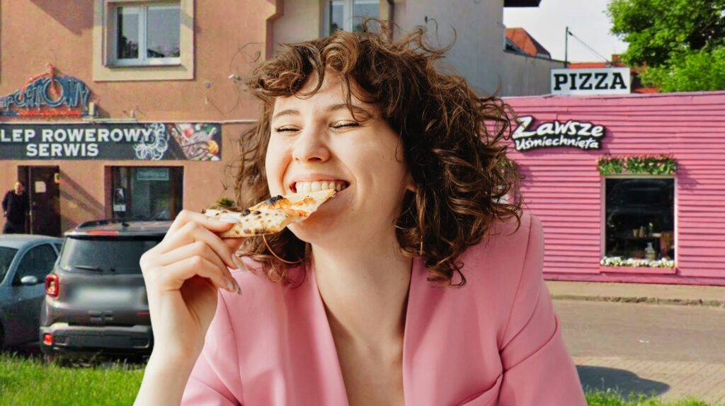 Kobieta w różowej marynarce uśmiecha się, jedząc pizzę.