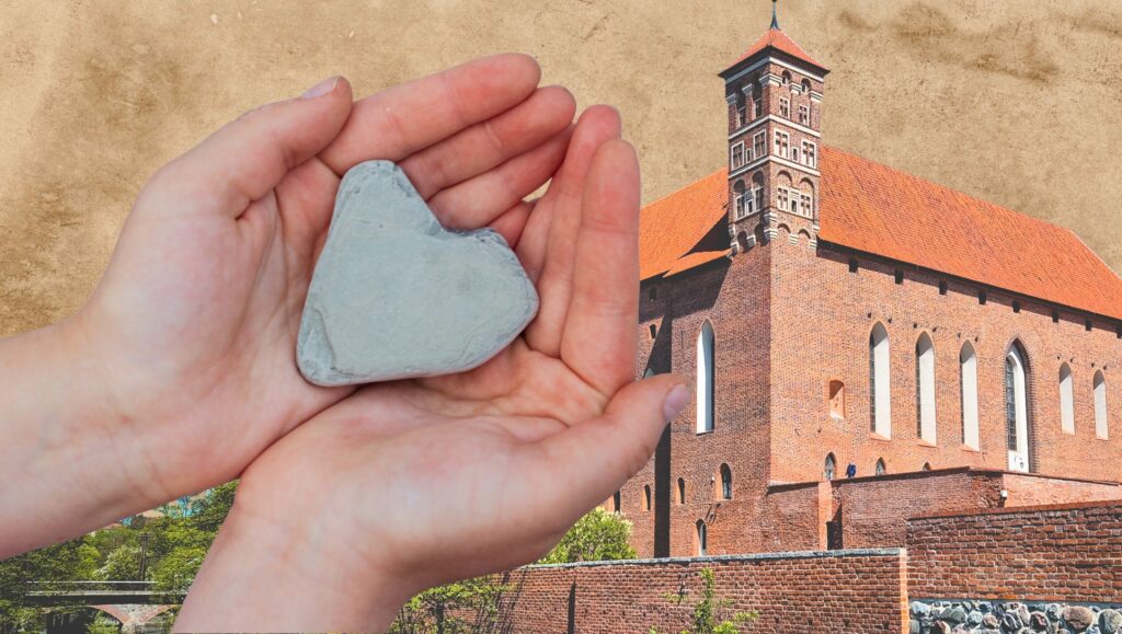 Dwie ręce trzymają szary kamień w kształcie serca, w tle budynek z czerwonym dachem.