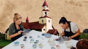 Dwie kobiety malują naczynia ceramiczne przy stole.