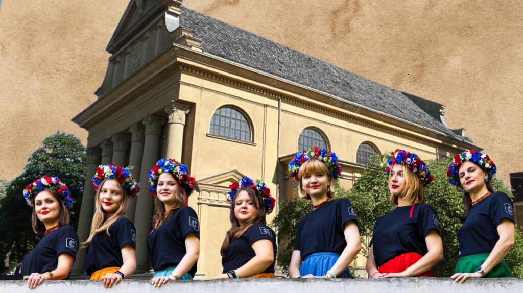 Grupa kobiet w tradycyjnych strojach z kwiatowymi wiankami przy budynku.