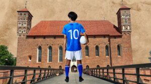 Młody piłkarz w niebieskiej koszulce z numerem 10 trzyma piłkę przed średniowiecznym zamkiem.