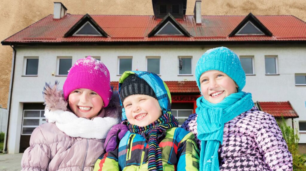 Trójka dzieci w zimowych kurtkach i nakryciach głowy przed budynkiem.