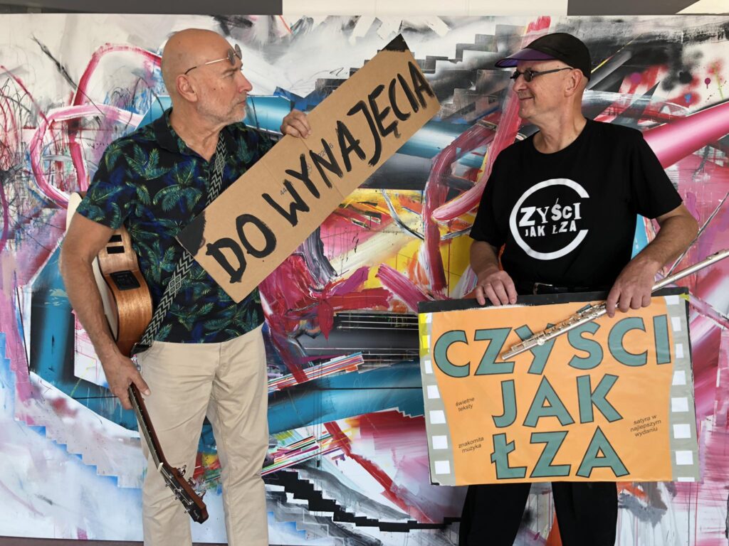 Dwóch mężczyzn trzymających plakaty w artystycznym otoczeniu.