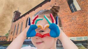 Dziecko z kolorowymi dłońmi formującym serce na tle ceglanej ściany.