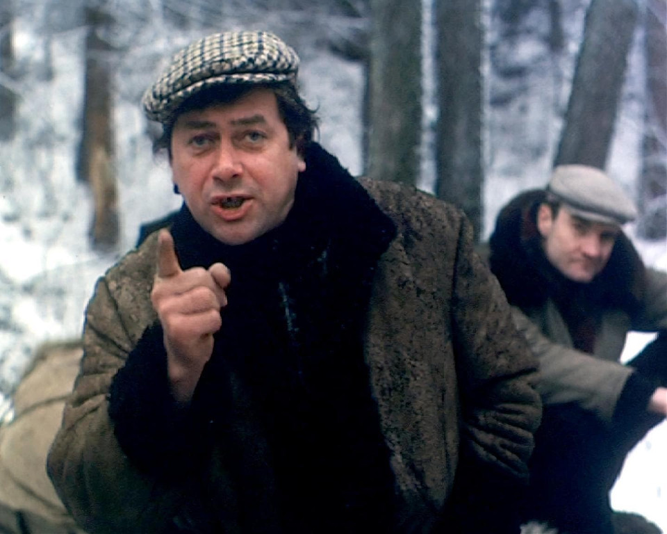 Mężczyzna w futrzanej kurtce z kapeluszem na tle lasu zimą.