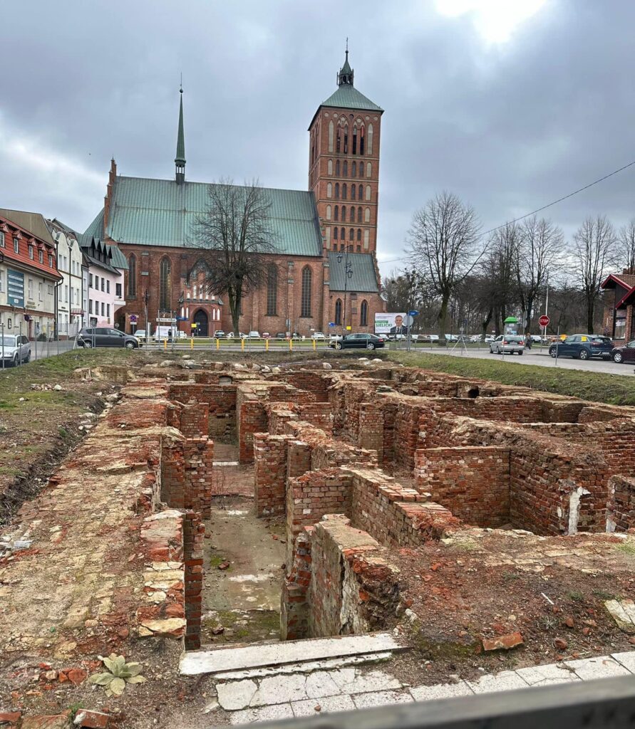 Ruiny budowli z cegły i kościół w tle.