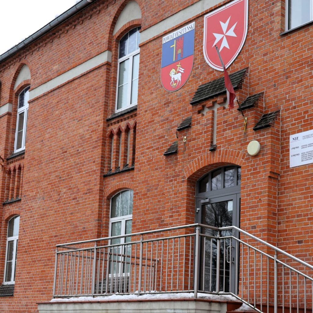 Czerwony budynek z tarasem i herbami.