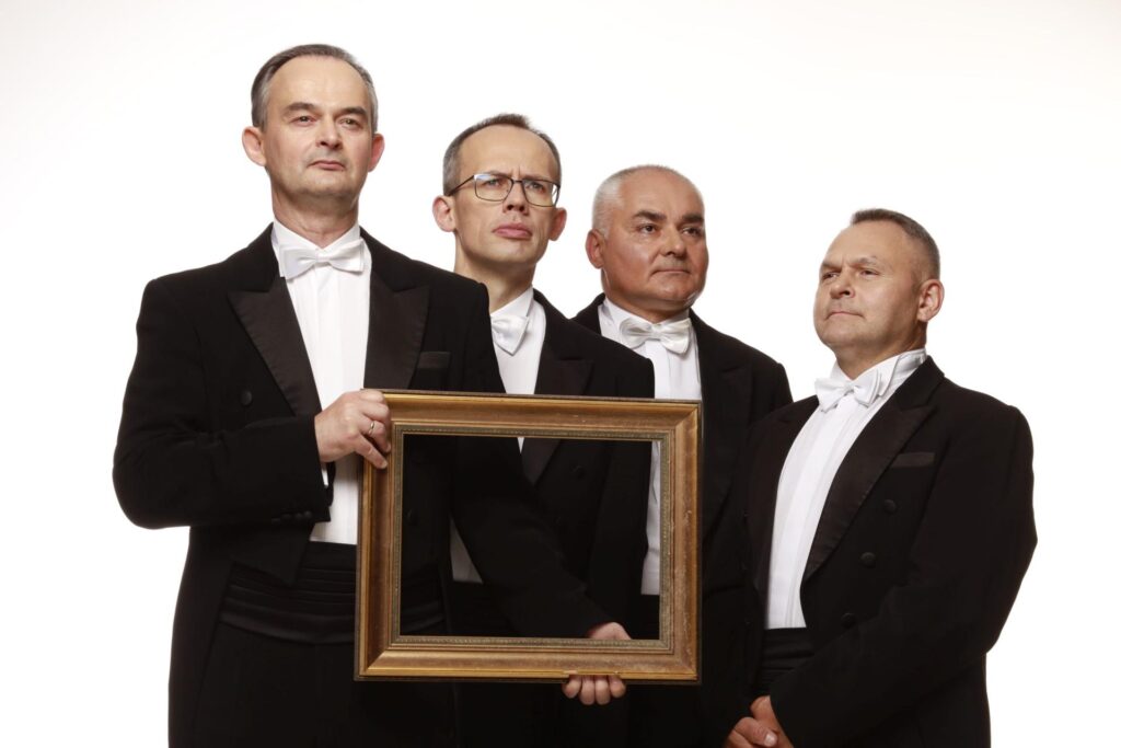 Czterech mężczyzn w smokingach trzyma prostokątną ramkę.