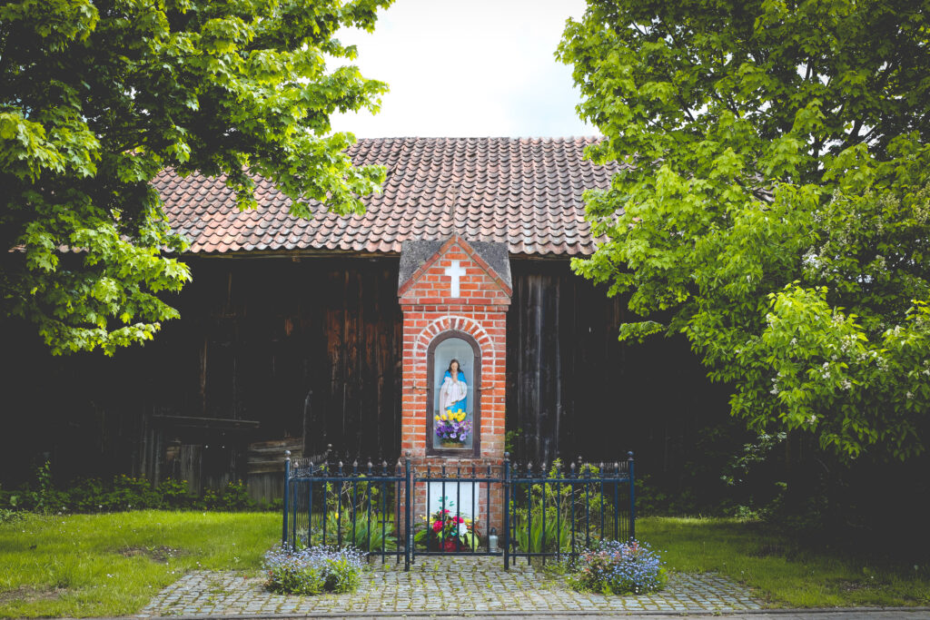 Kapliczka z rzeźbą Marii przed drewnianym budynkiem.