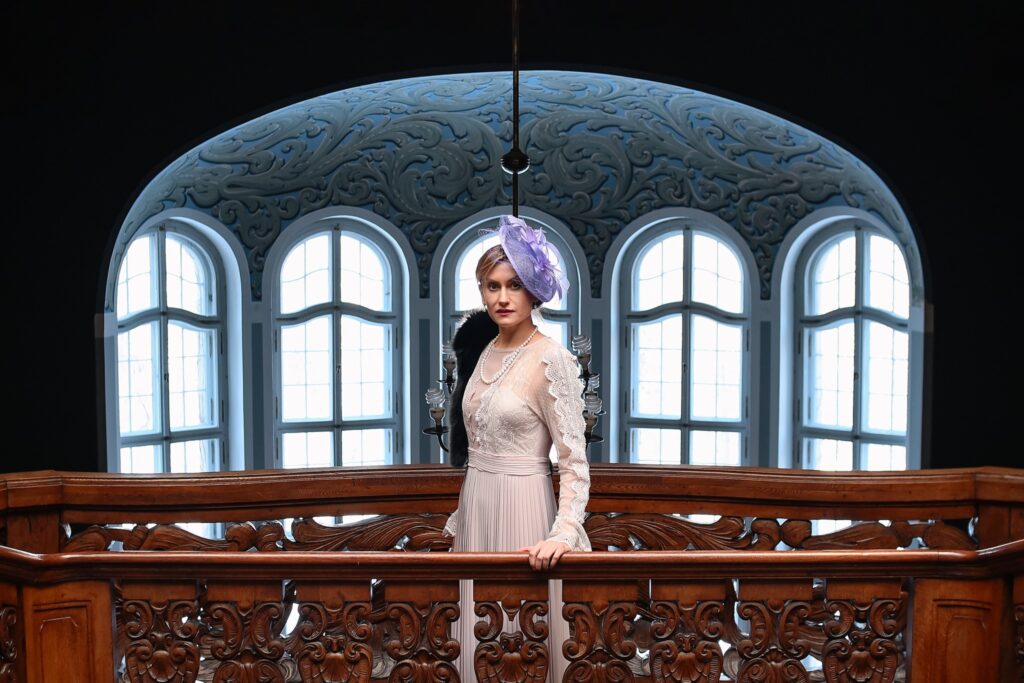 Kobieta w eleganckiej sukni stoi na balkonie z rzeźbioną balustradą.