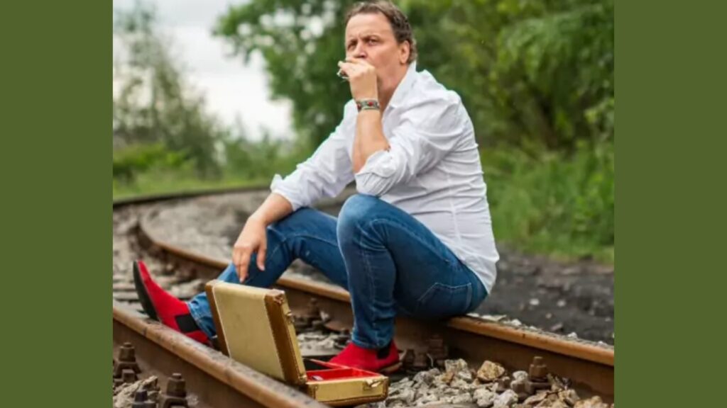 Mężczyzna w białej koszuli siedzący na torach kolejowych i grający na harmonijce.