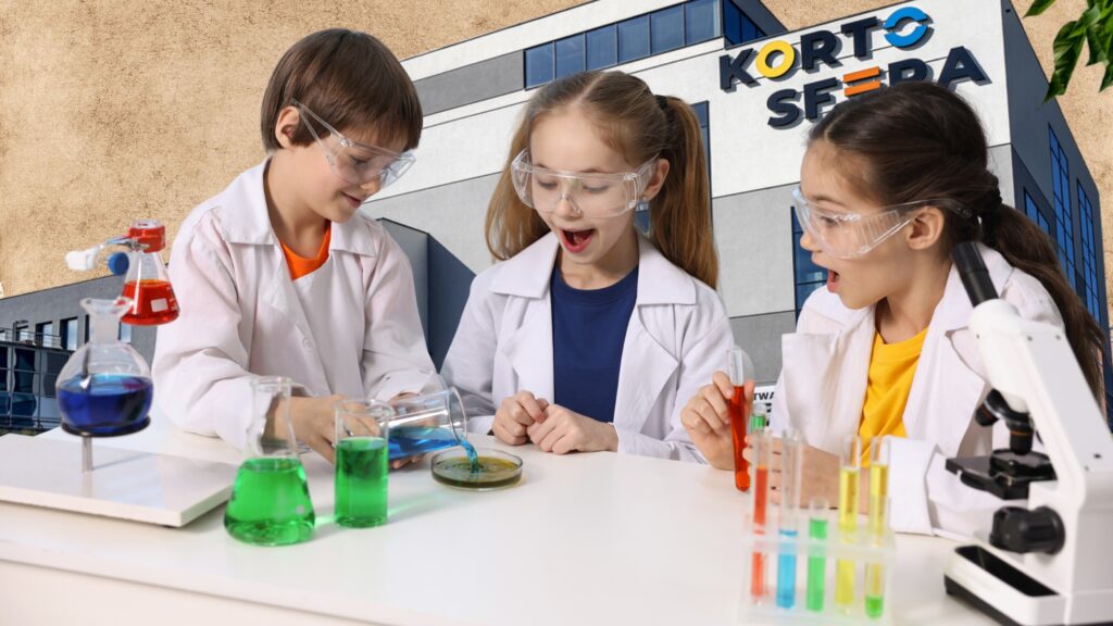 Troje dzieci w białych fartuchach i okularach ochronnych wykonujących eksperymenty chemiczne na tle budynku z napisem „KORTOSFERA”