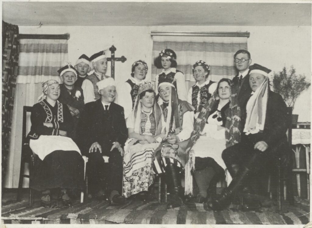 Grupa osób w tradycyjnych strojach ludowych na tle wnętrza.