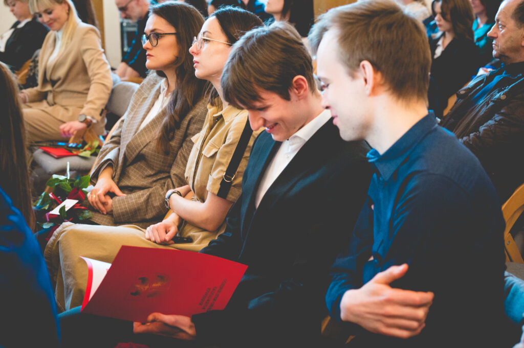Grupa młodych ludzi siedzi na sali, uśmiechając się i trzymając dokumenty.