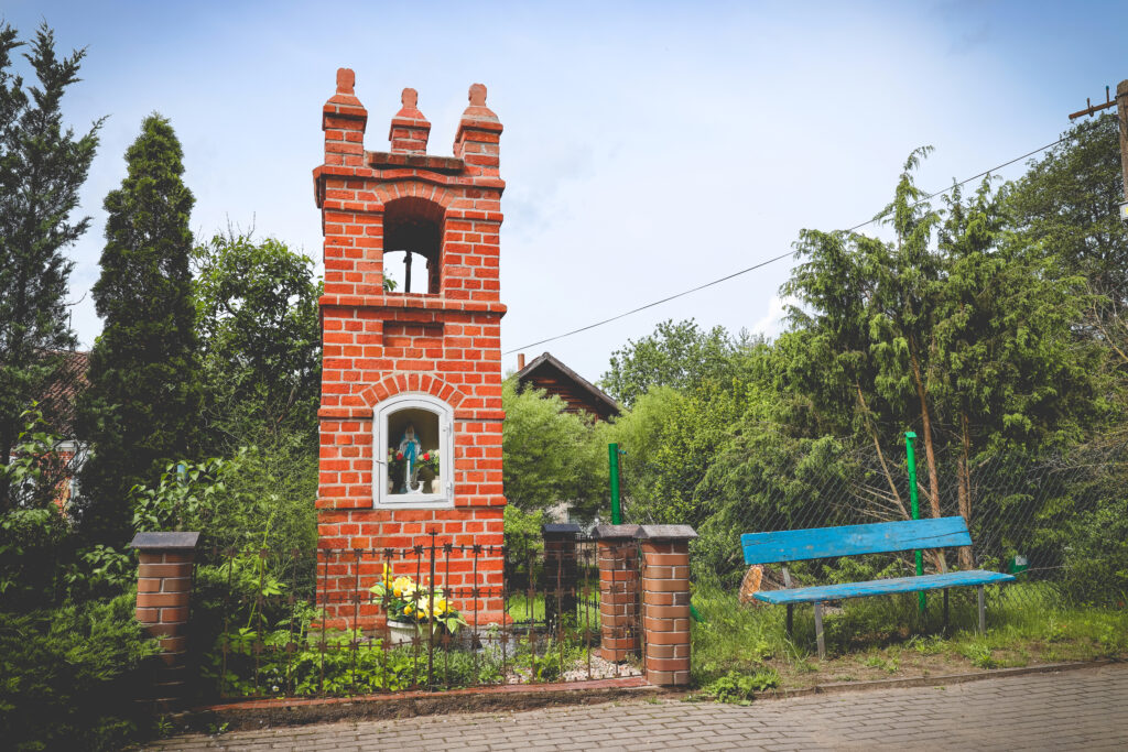 Czerwona ceglana kapliczka z figurą i ławeczka na tle zieleni.