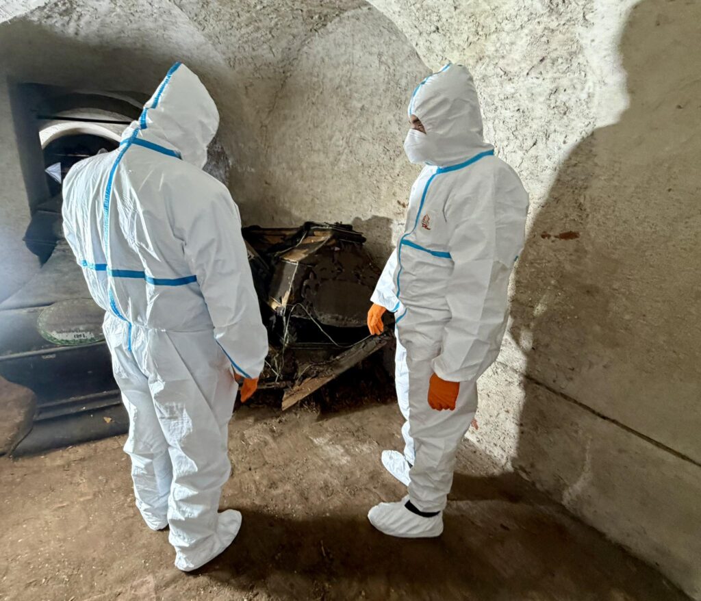 Dwie osoby w kombinezonach ochronnych badają przedmiot w zamkniętej przestrzeni.