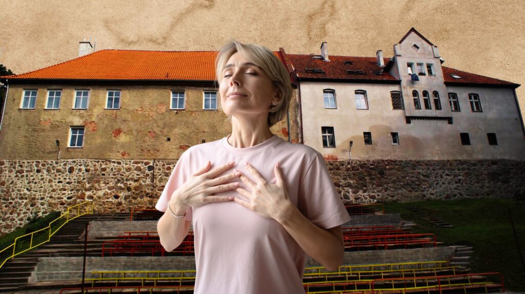 Kobieta w różowej koszulce stoi z zamkniętymi oczami przed zabytkowym budynkiem.