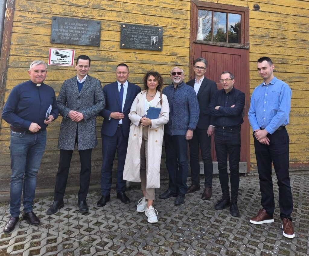 Grupa osób stojąca przed drewnianym budynkiem.