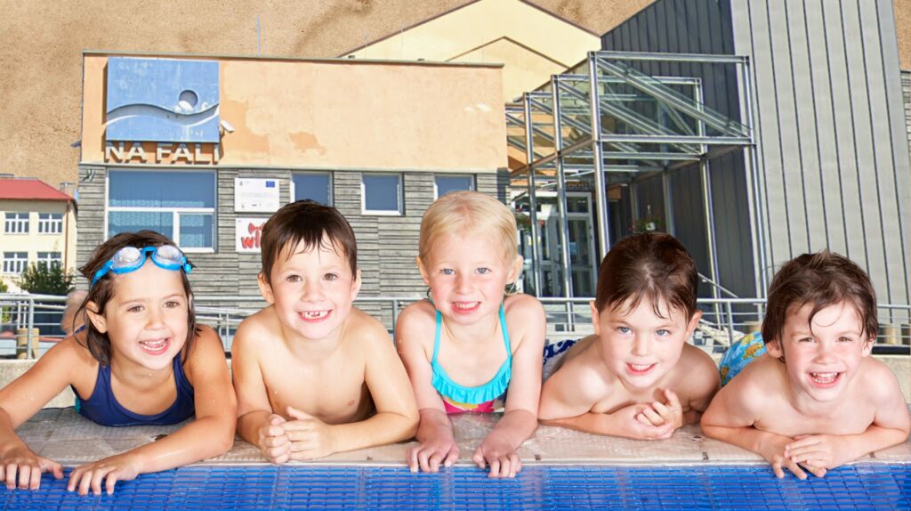 Pięcioro dzieci w kąpielówkach przy basenie, uśmiechniętych.