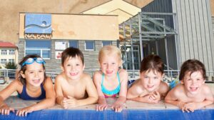 Pięcioro dzieci w kąpielówkach przy basenie, uśmiechniętych.