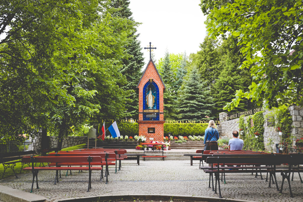 Kapliczka z figurą Matki Boskiej otoczona zielenią.