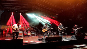 Pięcioosobowy zespół muzyczny grający koncert na dużej scenie plenerowej, oświetlonej dynamicznymi czerwonymi i białymi reflektorami.