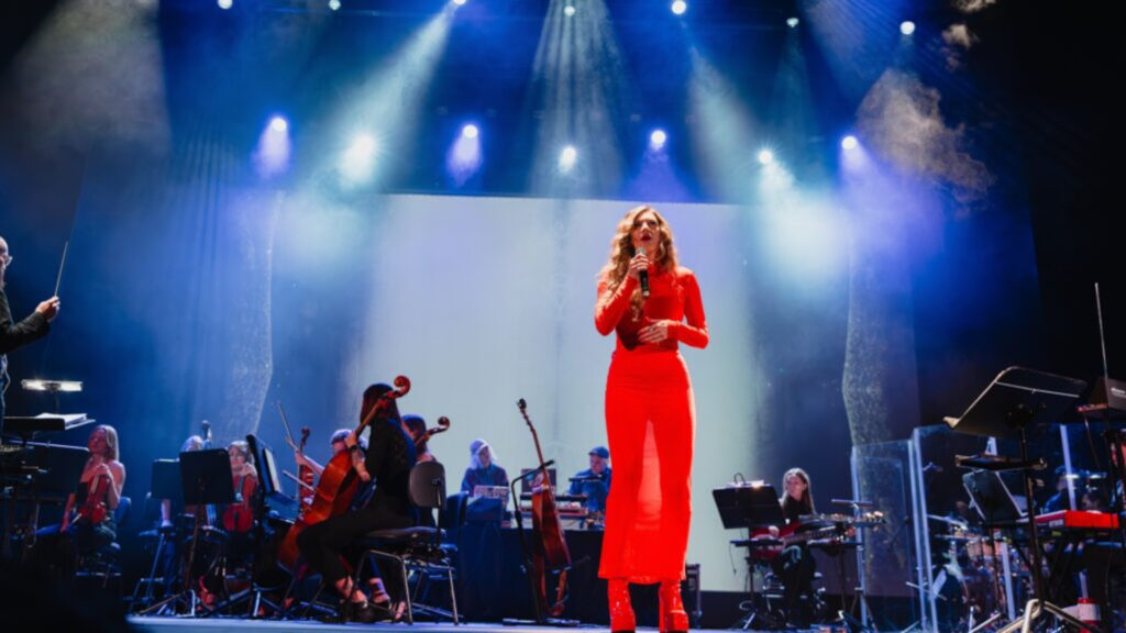 Wokalistka w jaskrawej, czerwonej sukni śpiewająca na scenie filharmonii w towarzystwie orkiestry, oświetlona mocnymi niebiesko-białymi reflektorami.