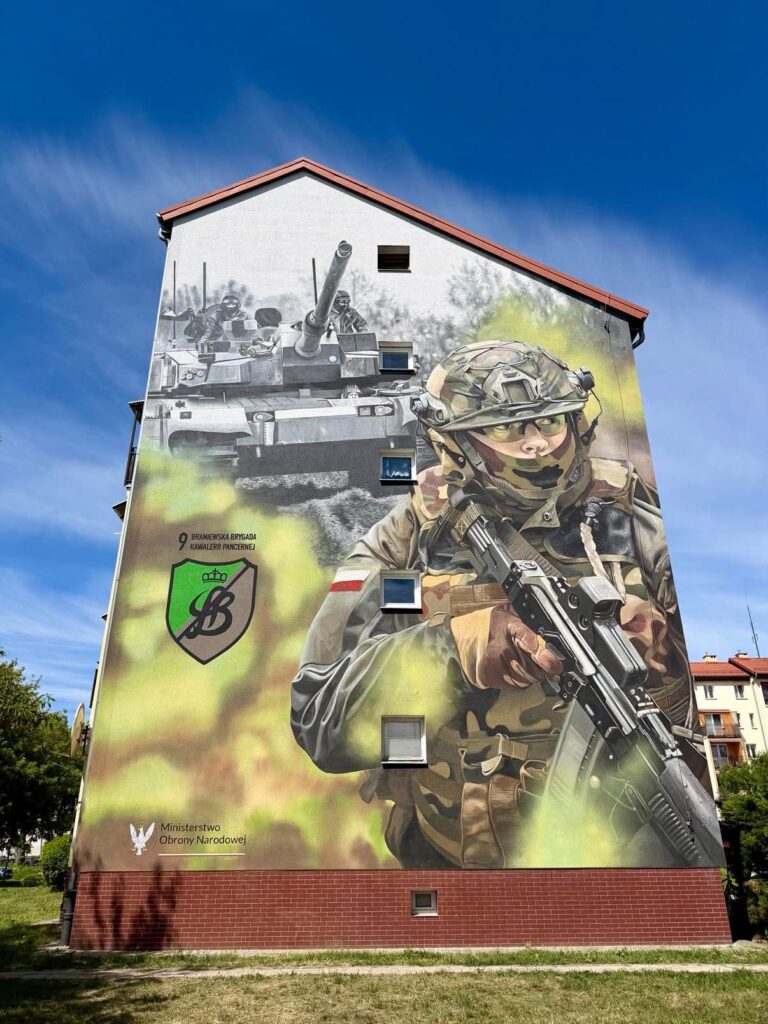Wielki mural przedstawiający żołnierza z bronią i czołg w tle na budynku.