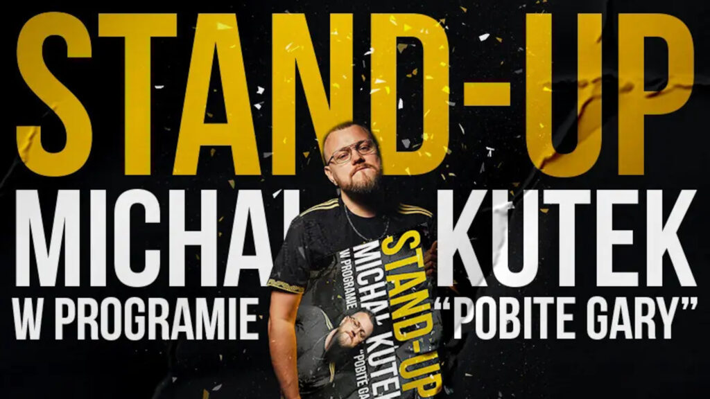 Komik Michał Kutek w okularach, stojący na tle czarnego plakatu z wielkim żółtym napisem „STAND-UP” i trzymający w rękach mniejszą wersję tego samego plakatu.