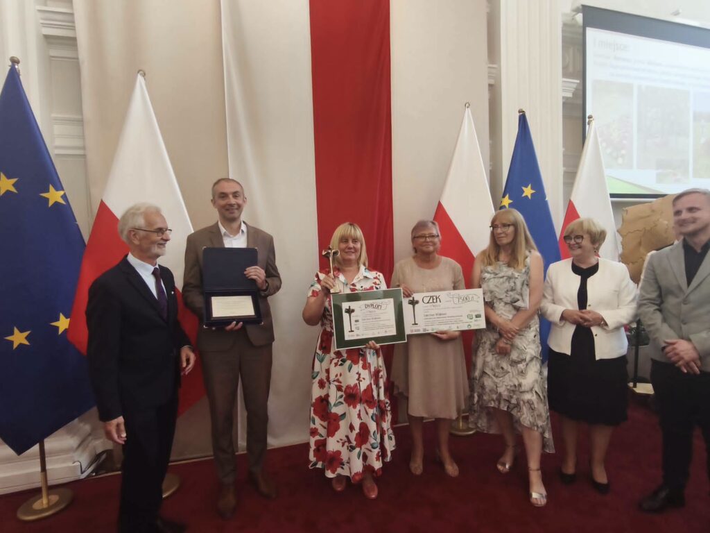 Grupa osób na uroczystości z dyplomami w tle flagi polskiej i unijnej.